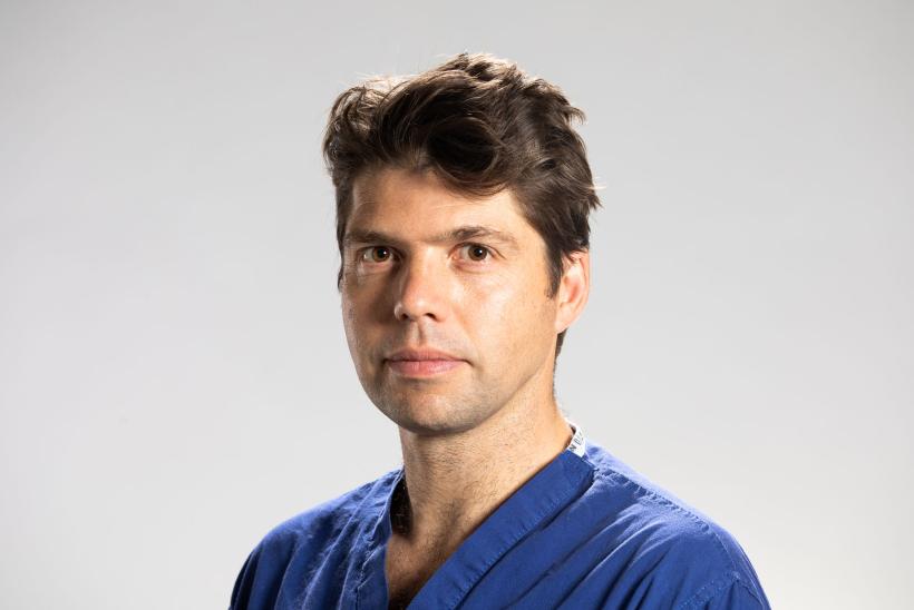 صورة الطبيب الجراح أليكس وولارد، مرتدياً البزة الجراحية الزرقاء وواقفاً أمام خلفية بيضاء تميل نحو الرمادية تدريجياً من الأسفل للأعلى.