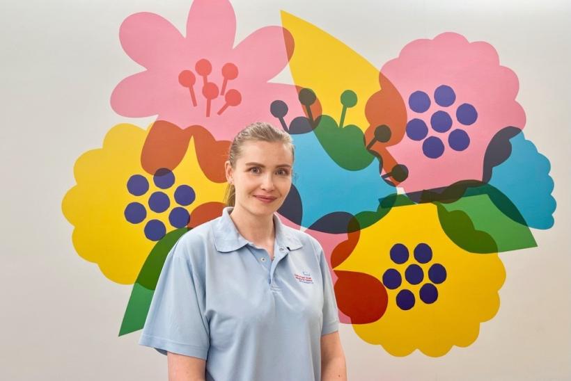 تظهر السيدة إيديل دو بويتلير مرتديةً قميصاً سماوي وخلفها حائط عليه زهور بألوان وردية، زرقاء، صفراء، وحمراء.