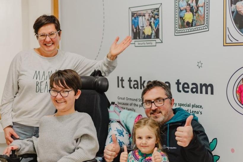 A family of 4 pose in front of the Family Wall where they're also featured
