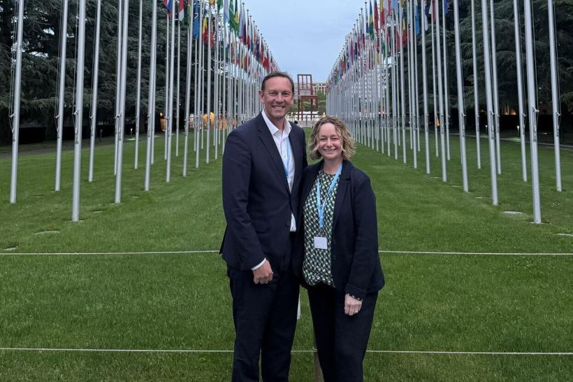 Lyn Shields (right) and Chris Rockenbach (left) are stood in between the flag poles of different nations with green grass under them at the European headquarters of the U.N. in Geneva, Switzerland.