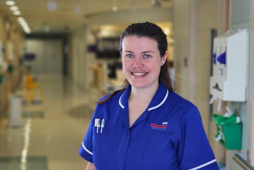 Jasmin Dowdall who is the Caterpillar/Outpatients Ward Manager stood in a hallway in a ward clad in blue uniform and smiling at the camera.