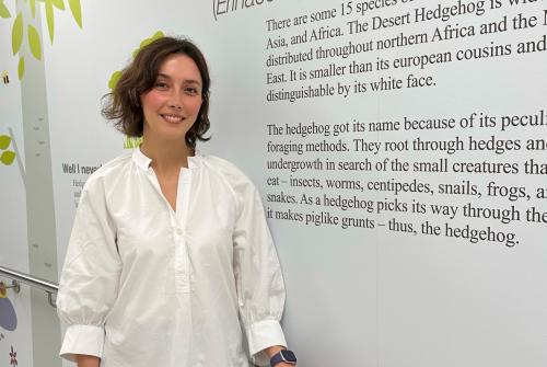 Dr Yasmine Olabi, Specialist Paediatric Clinical Psychologist, is stood smiling at the camera on the Bumblebee Ward in a hallway with writing on the wall next to her.