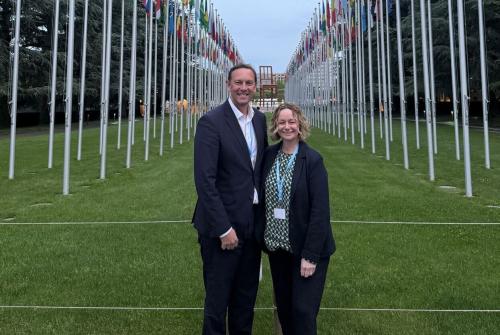 Lyn Shields (right) and Chris Rockenbach (left) are stood in between the flag poles of different nations with green grass under them at the European headquarters of the U.N. in Geneva, Switzerland.