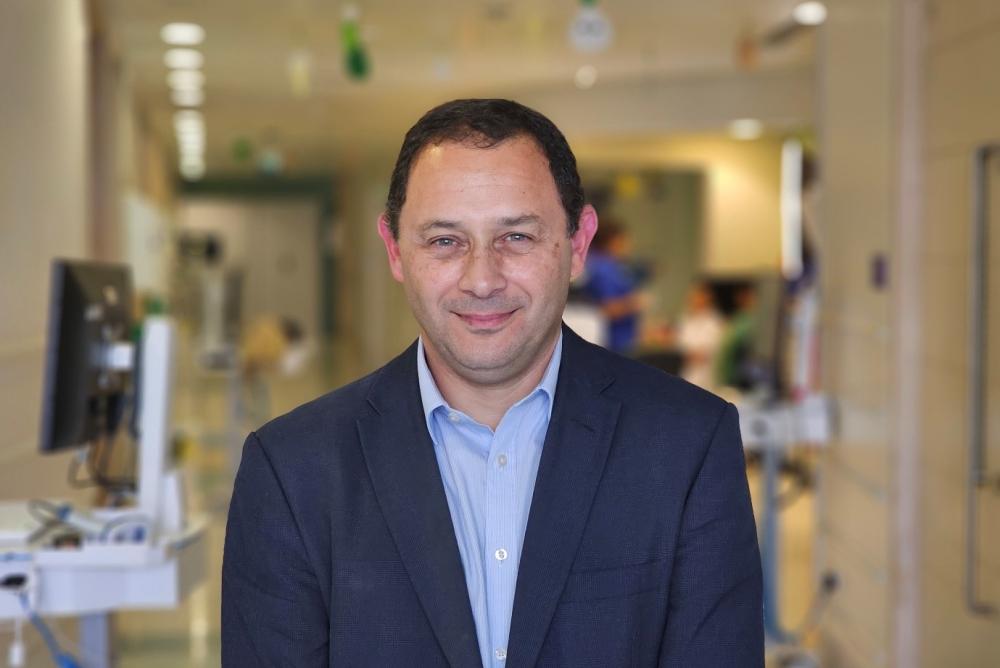 الطبيب الجراح كريستيان أكويلينا يبتسم للكاميرا في أحد أجنحة مستشفى جريت أورموند ستريت مرتدياً بدلة زرقاء داكنة وقميصاً أزرق.